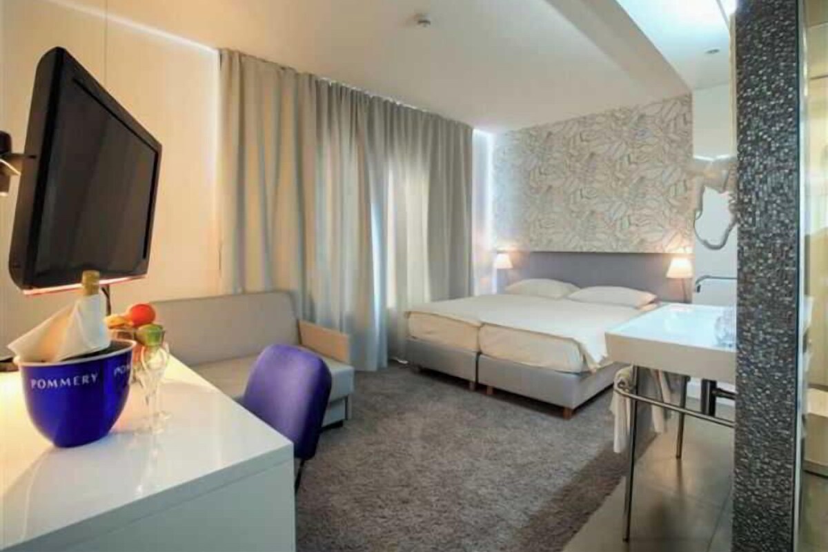 A hotel room with a bed, a desk with a computer, a chair, a TV, and a bathroom with a sink and mirror.