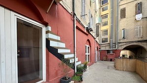 Un callejón estrecho con un edificio rojo a la izquierda, una cerca de madera a la derecha y un pequeño jardín con plantas en macetas.