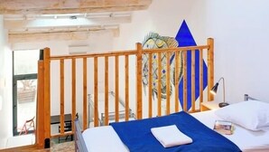 Un dormitorio con techo inclinado, tragaluz, cama con manta azul, cabecero de madera y pared con un cuadro geométrico.