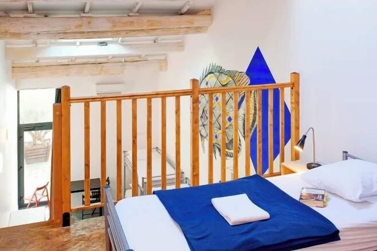 Une chambre avec un plafond en pente, un puits de lumière, un lit recouvert d’une couverture bleue, une tête de lit en bois et un mur orné d’un tableau aux motifs géométriques.