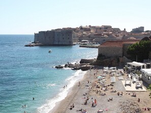 A coastal cityscape with a prominent historic building, a beach with sun loungers, and a clear view of the sea.
