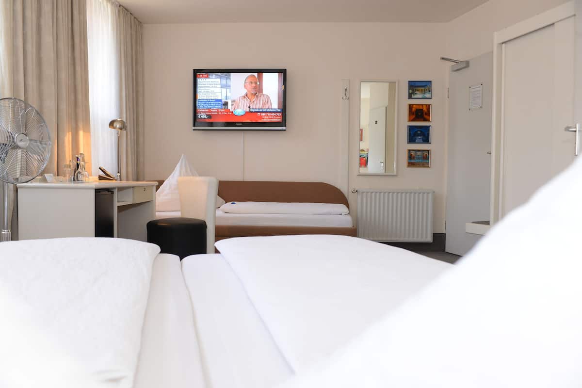 A hotel room with two beds, a TV on the wall, a desk with a lamp, a fan, and a window with curtains.