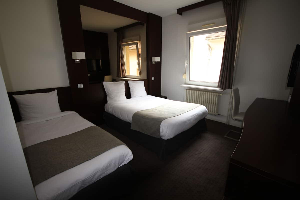 A hotel room with two single beds, a large window with curtains, a radiator, and a television on a stand.