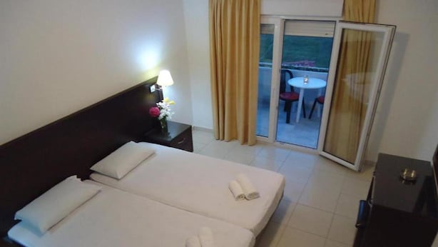 Quarto de hotel com duas camas, uma mesinha de cabeceira com abajur, uma pequena mesa com um vaso de flores e uma porta de vidro deslizante que dá para uma área externa com mesa e cadeiras.