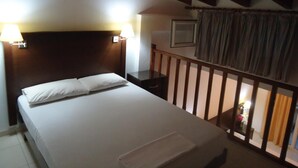 Quarto de hotel com cama, duas luminárias de cabeceira, painel de cabeceira de madeira, mesa de cabeceira e uma pequena mesa com abajur, além de janela com cortinas.