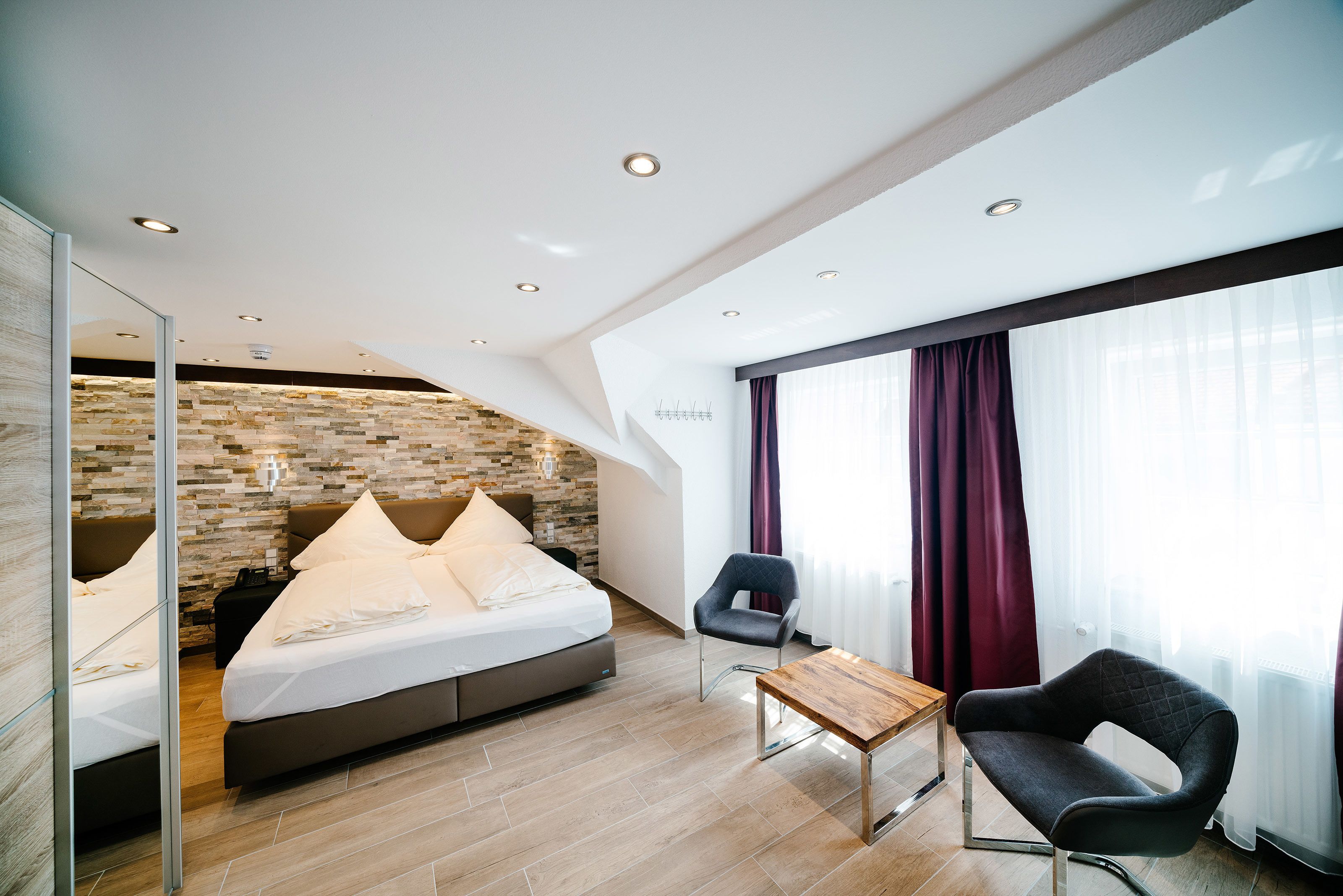 A modern hotel room with a stone accent wall, a bed with white linens, a wooden table, a black armchair, and a large window with curtains.