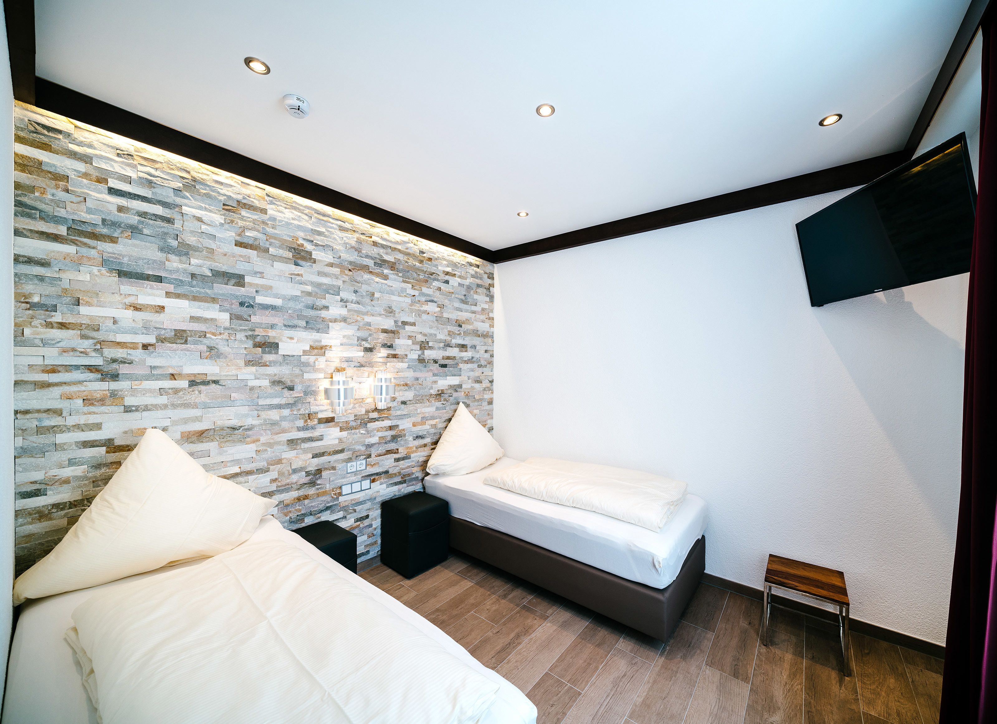 Une chambre moderne avec un mur en pierre, un lit, une table de chevet, un téléviseur fixé au mur et des spots encastrés au plafond.