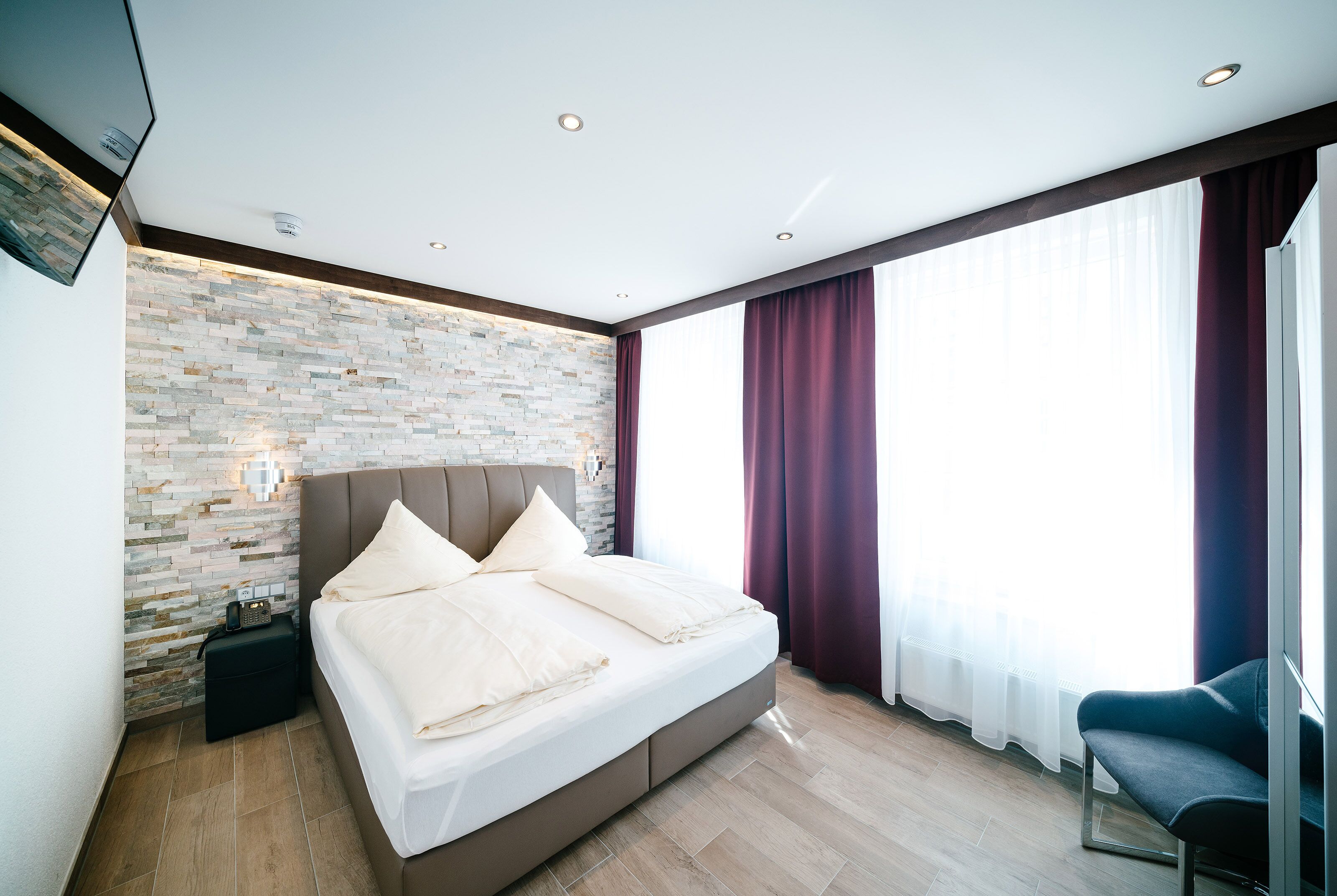 A modern bedroom with a stone wall, a large bed, a bedside table, and a view of the outdoors through sheer curtains.