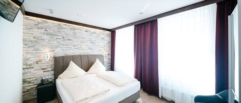 Un dormitorio moderno con una pared de piedra, una cama grande, una mesita de noche y vistas al exterior a través de cortinas de gasa.