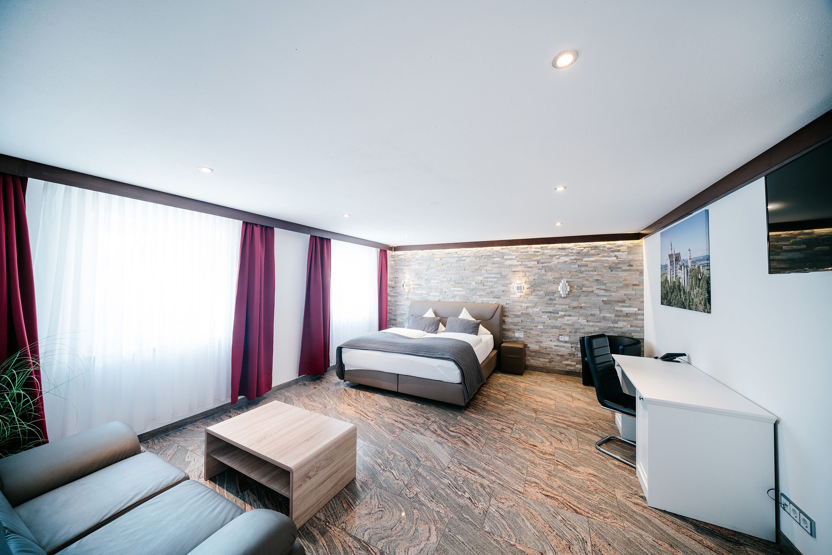 A modern hotel room with a bed, a desk, a sofa, and a large window with curtains.
