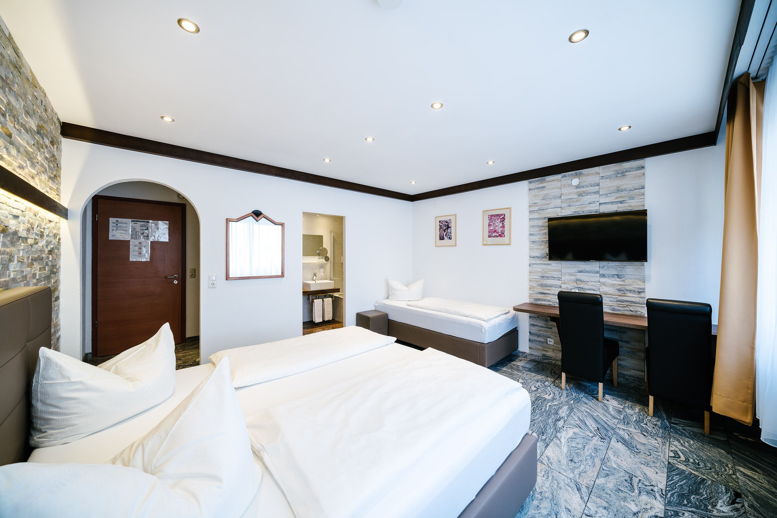 A hotel room with a large bed, a desk with two chairs, a TV mounted on the wall, and a bathroom visible through an archway.