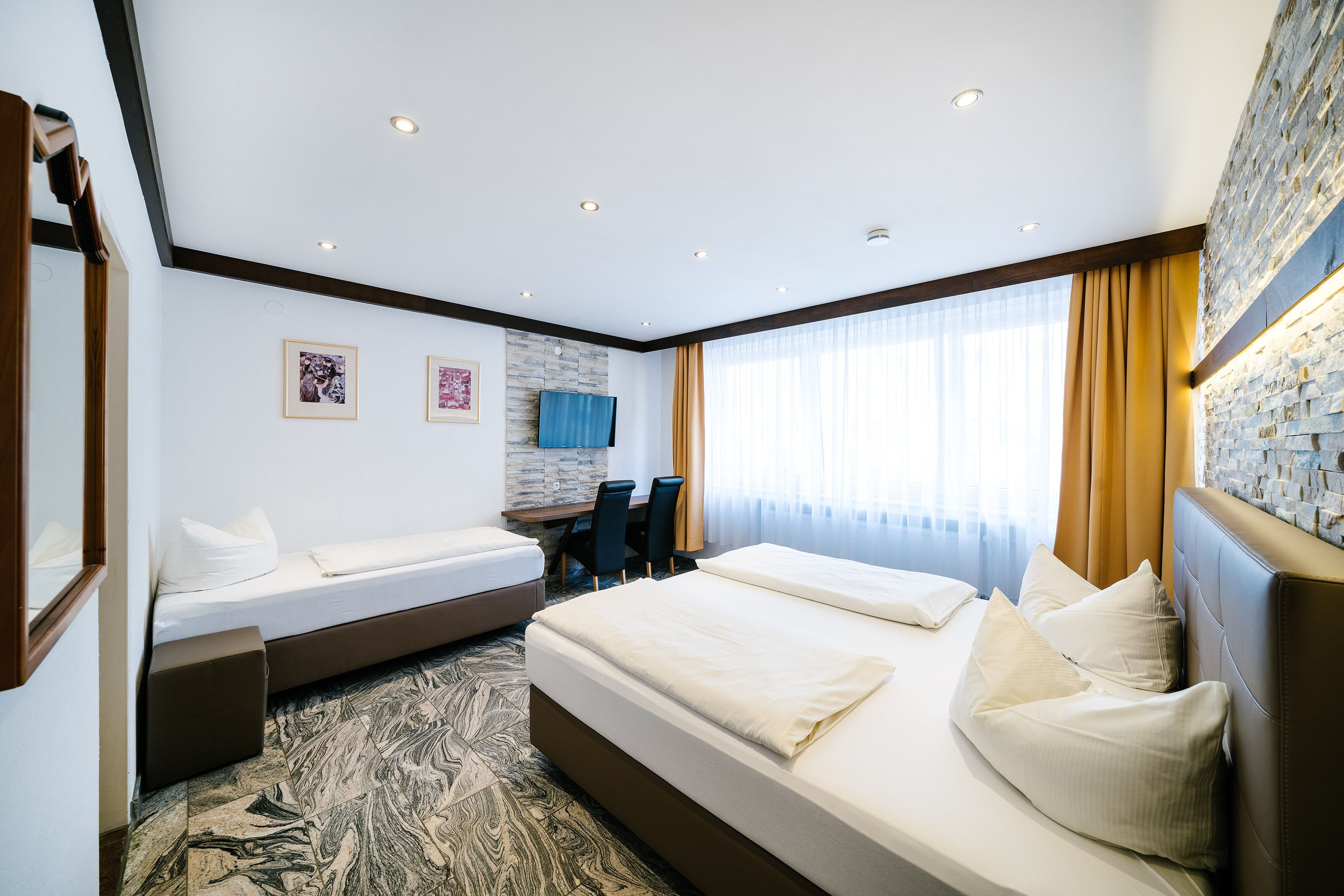 A hotel room with a large bed, a smaller bed, a desk with chairs, a television, and a stone accent wall.