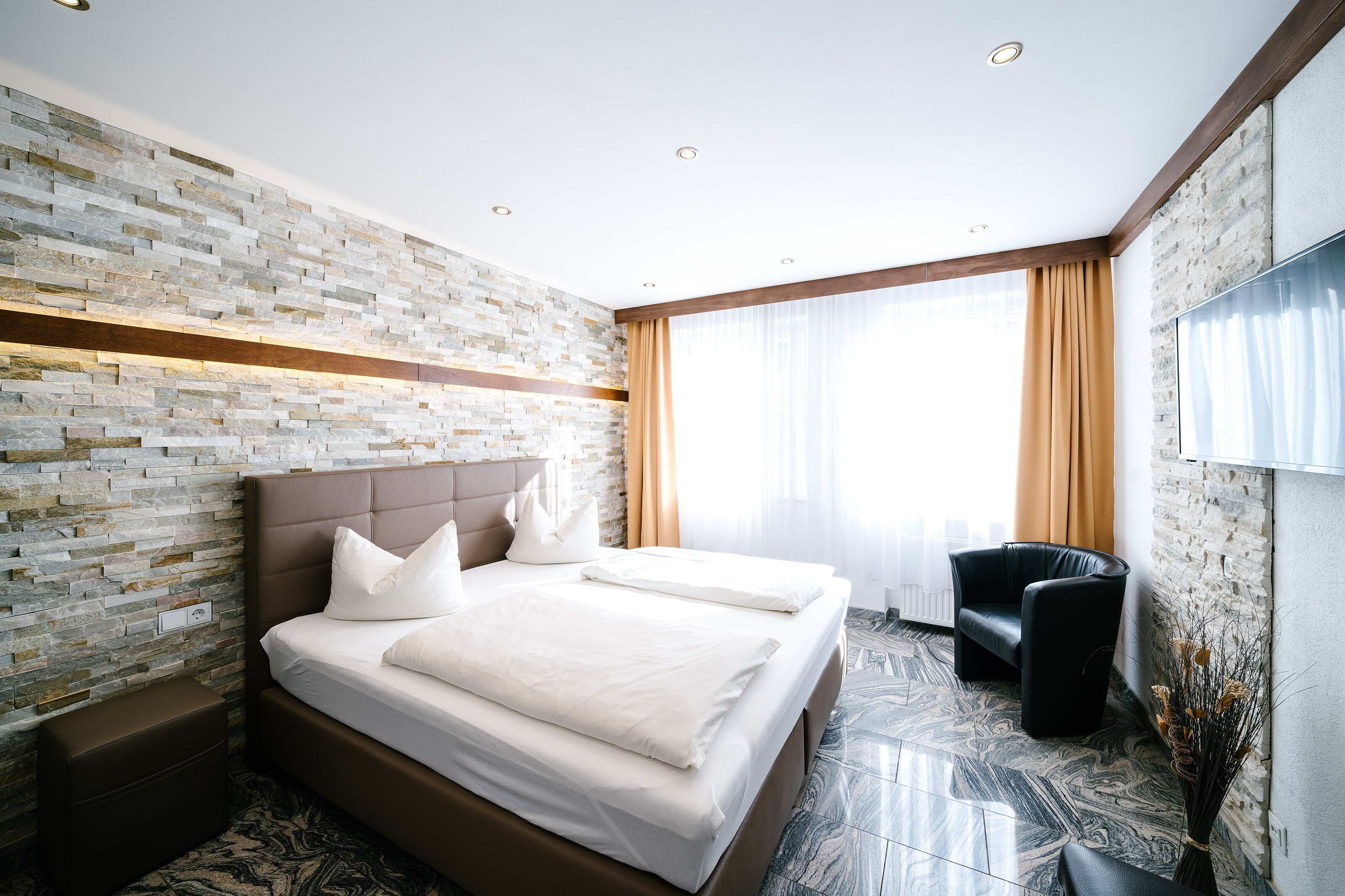 Una habitación de hotel moderna con pared de ladrillo, una cama grande con ropa blanca, un sillón de cuero negro y un televisor de pantalla plana en la pared.