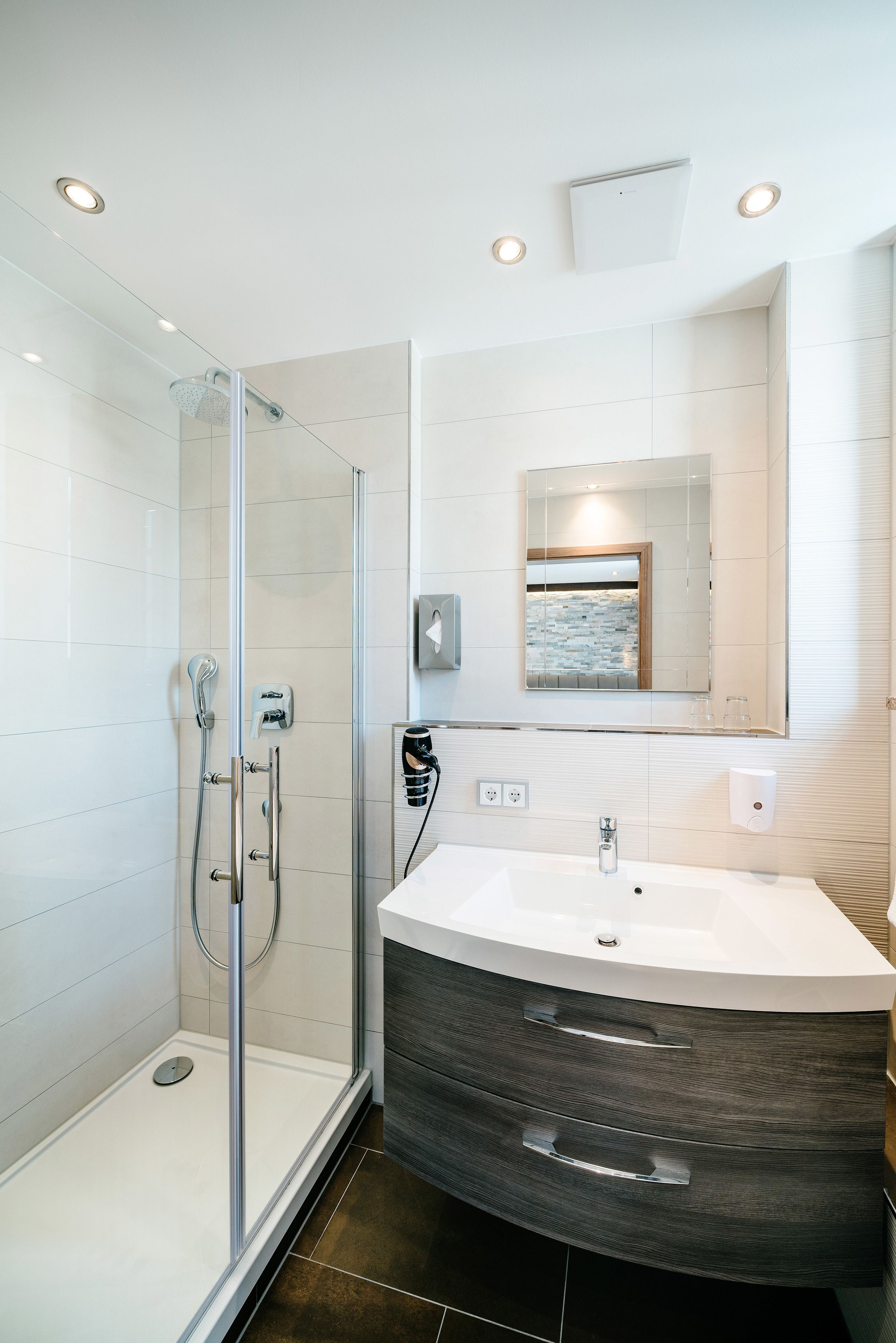 A modern bathroom with a glass-enclosed shower, a large vanity with a dark wood finish, and a mirror with a framed artwork above it.