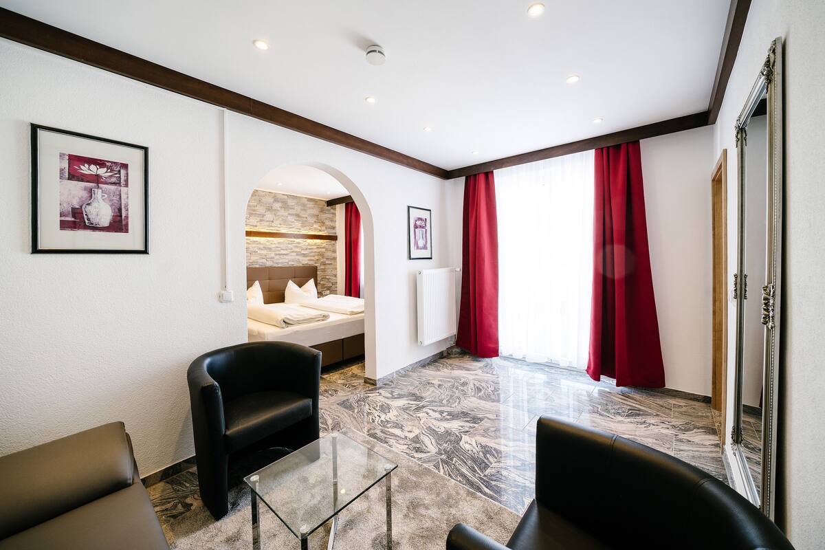 A hotel room with a bed, a seating area with chairs, a glass table, a fireplace, and red curtains.