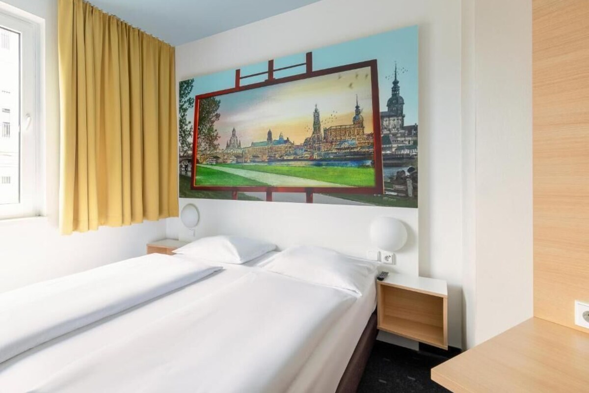 A hotel room with a bed, a nightstand, a wall-mounted TV, and a window with curtains.