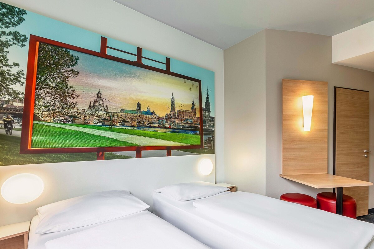 A hotel room with a bed, a nightstand, a wall-mounted light, and a painting of a cityscape.