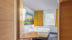 A hotel room with a bed, a desk, two red stools, a window with yellow curtains, and a painting of a cityscape. - B&B HOTEL Dresden-Messe (Dresden)