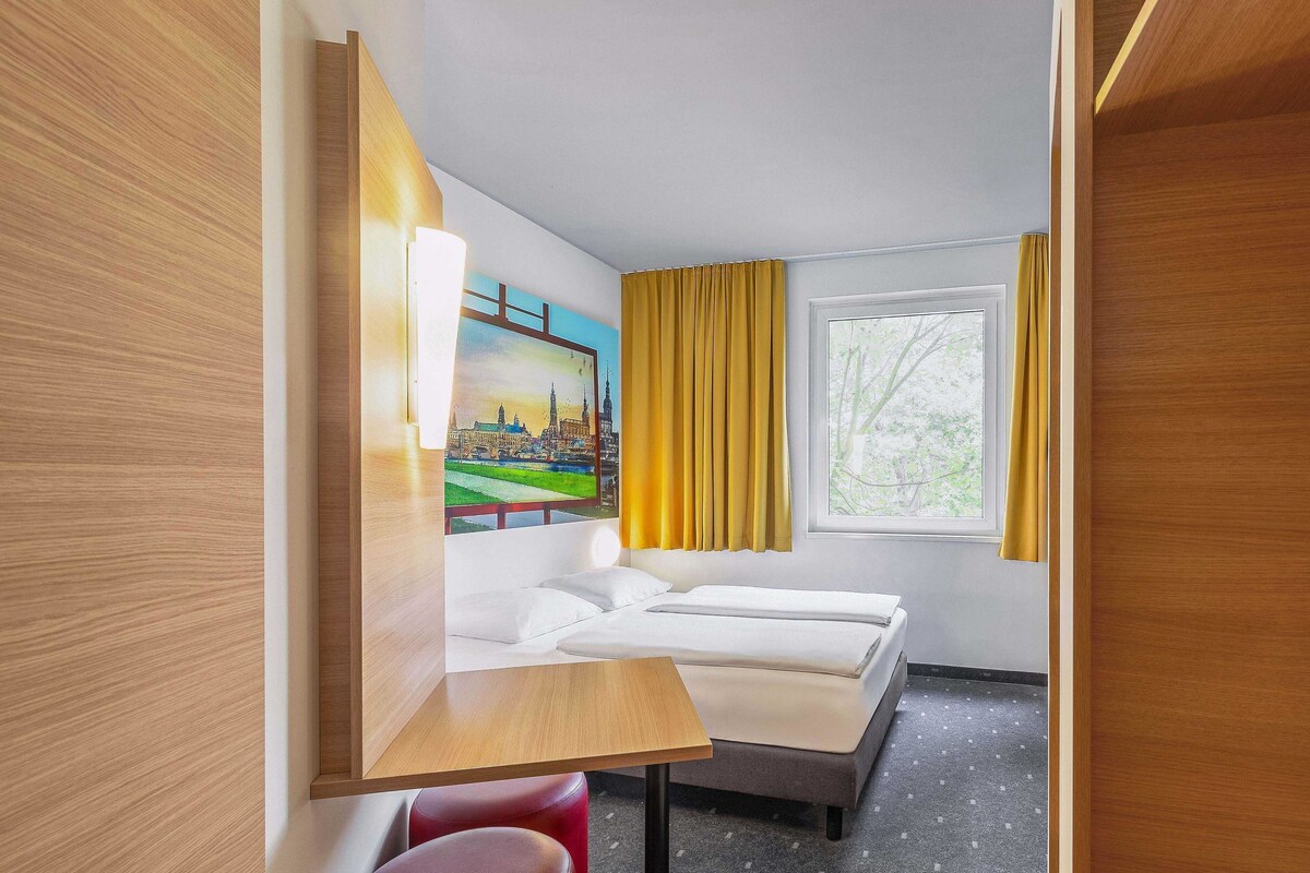A hotel room with a bed, a desk, two red stools, a window with yellow curtains, and a painting of a cityscape.