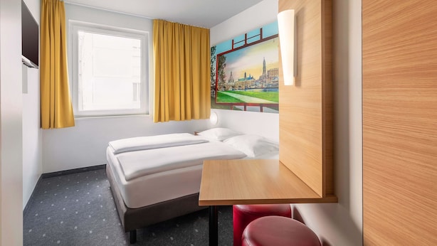 Une chambre d’hôtel avec un lit, un bureau, une chaise, un tableau représentant un paysage urbain et des rideaux.