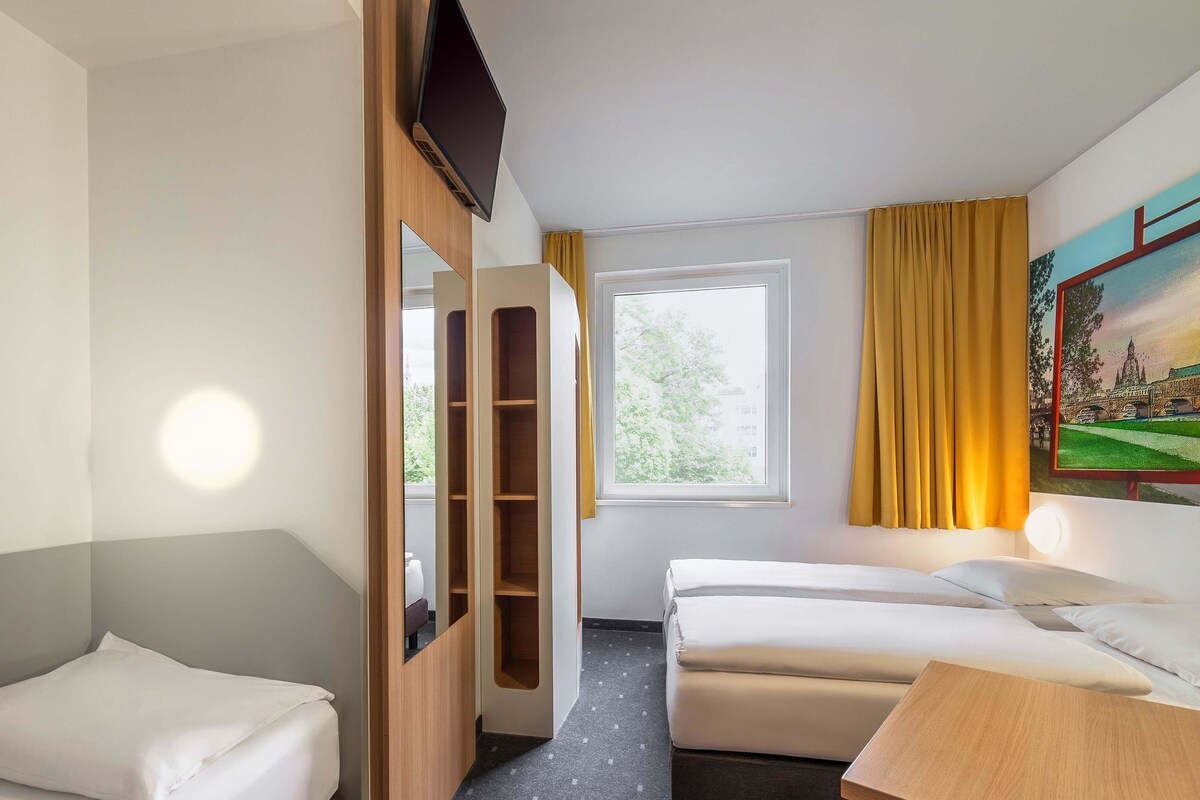 A hotel room with two single beds, a wooden wardrobe, a desk, a window with curtains, and a painting on the wall.