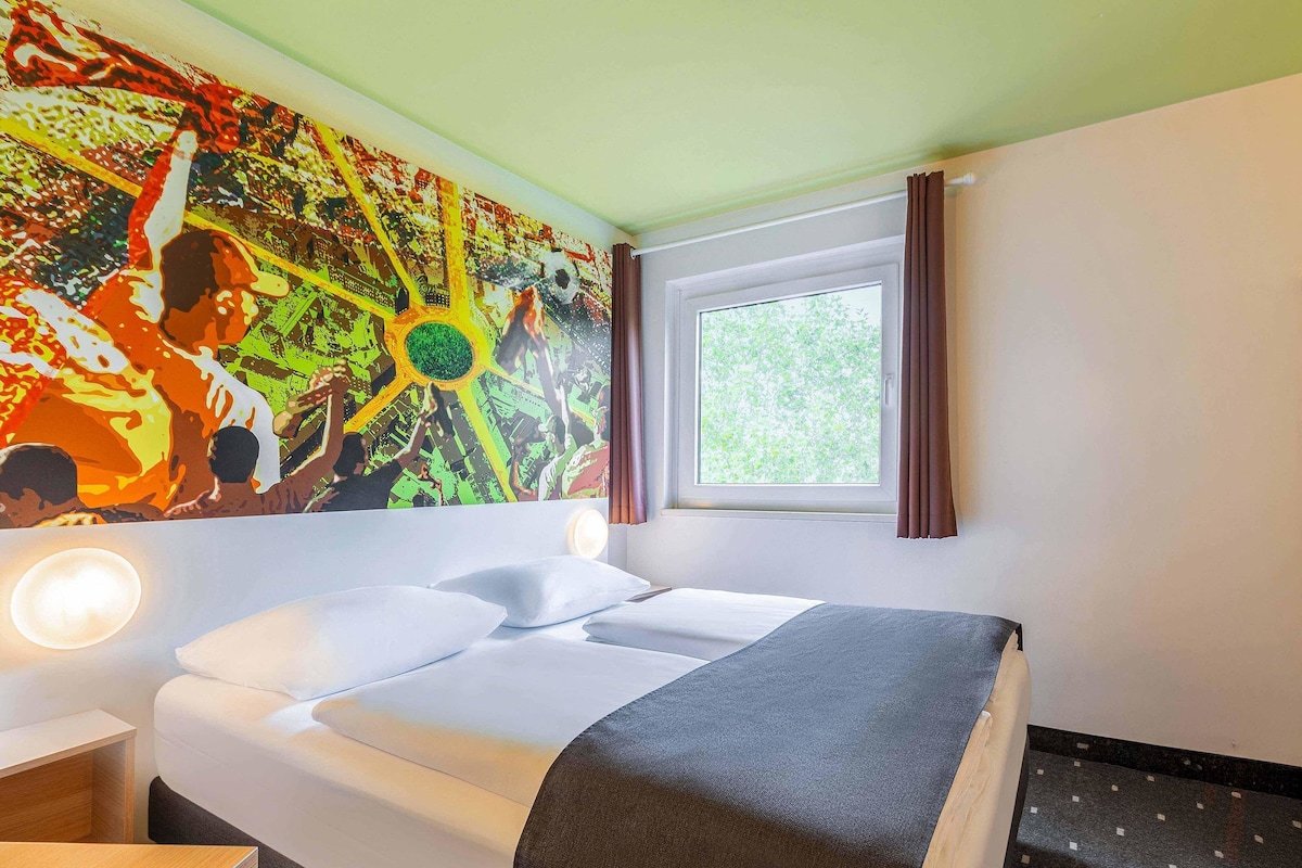 A hotel room with a bed, a nightstand, a window with curtains, and a colorful mural on the wall.