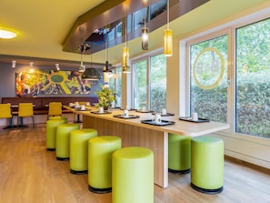 A modern restaurant with a long counter, yellow chairs, and green stools. - B&B Hotel Dortmund-Messe (Dortmund)