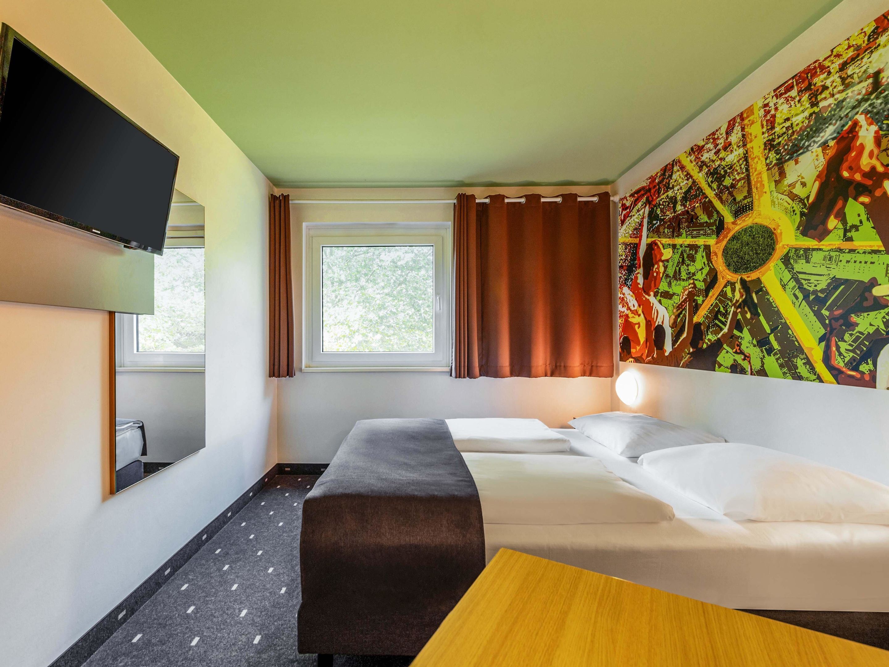 A hotel room with a bed, a desk, a TV, a window with curtains, and a large abstract painting on the wall.
