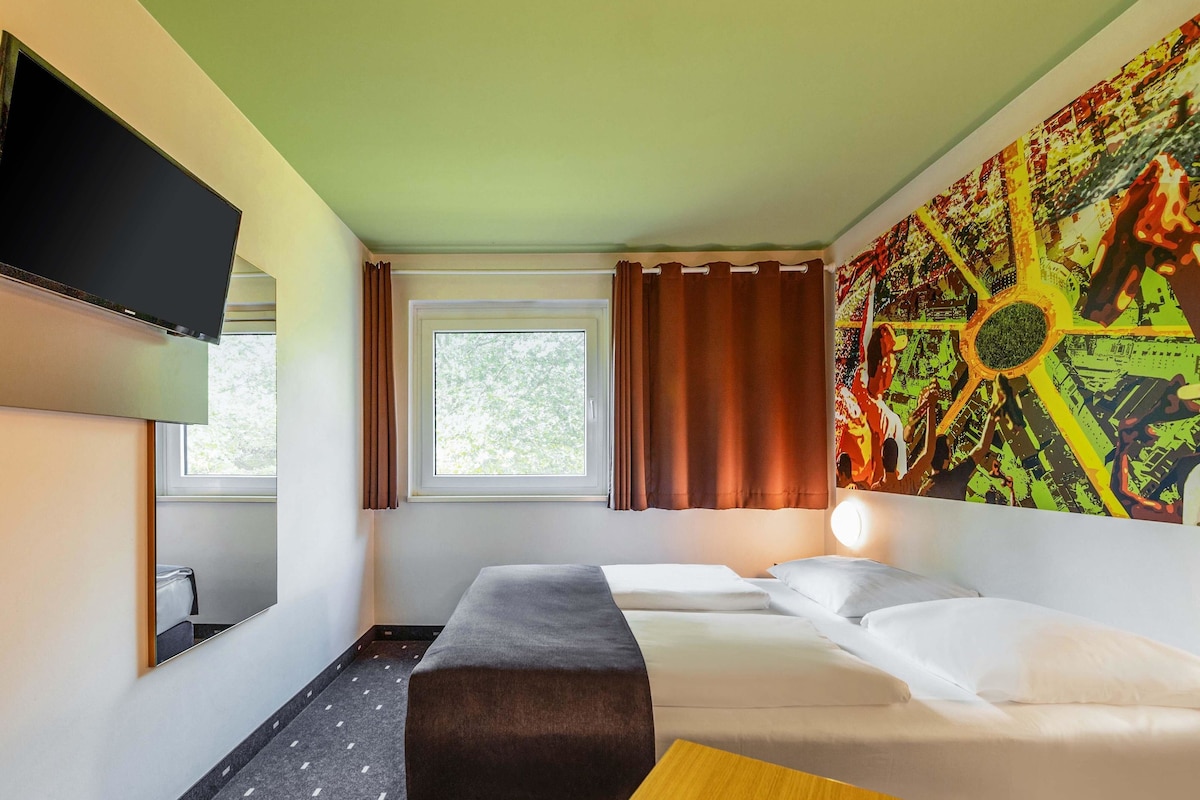 A hotel room with a bed, a desk, a TV, a window with curtains, and a large abstract painting on the wall.