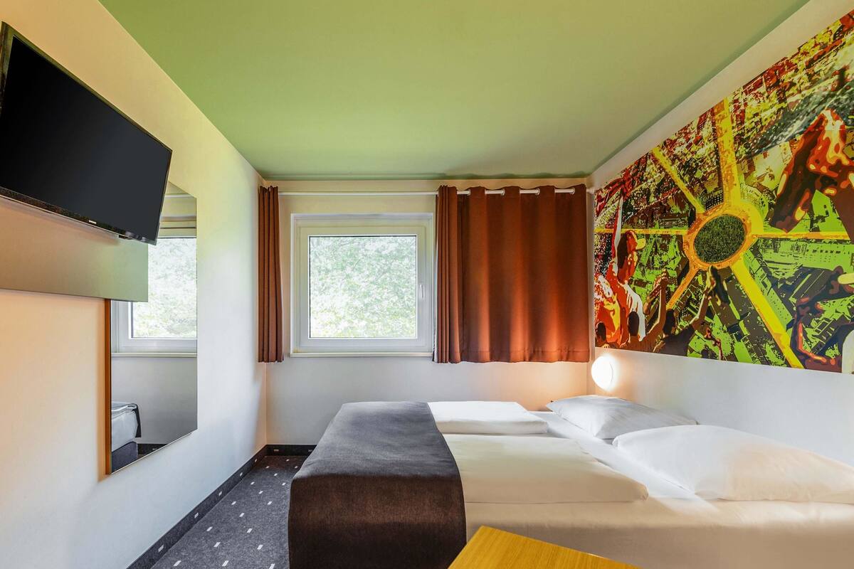 A hotel room with a bed, a desk, a TV, a window with curtains, and a large abstract painting on the wall.