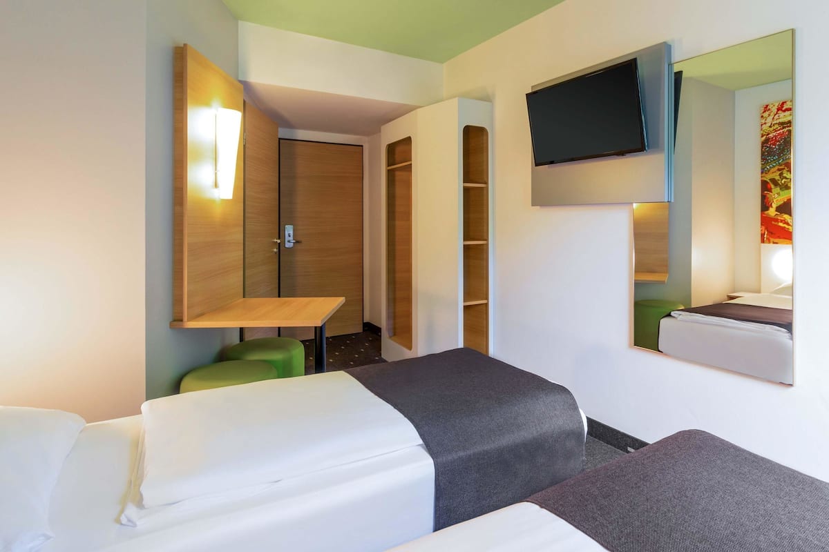 A hotel room with two beds, a TV mounted on the wall, a wooden wardrobe, and a small table with a green chair.