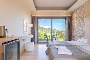 A hotel room with two beds, a minibar, a small desk, a balcony with a view of greenery, and a stone wall.