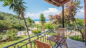 Un balcón con una mesa y una silla de madera, con vistas a un paisaje costero con árboles y el mar.