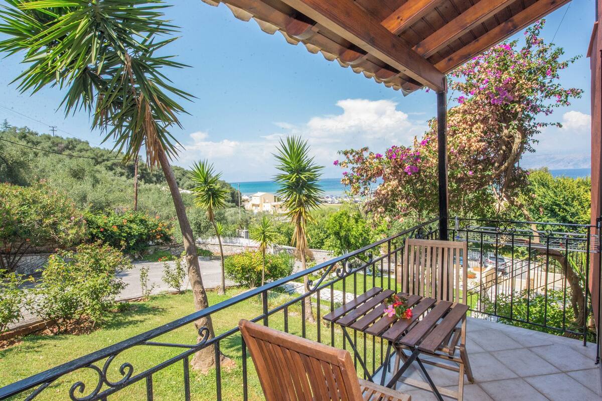 Ein Balkon mit Holztisch und Stuhl, der auf eine Küstenlandschaft mit Bäumen und Meerblick blickt.
