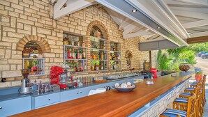 Un bar con pared de piedra, estantes con botellas, una máquina de café y sillas de mimbre.