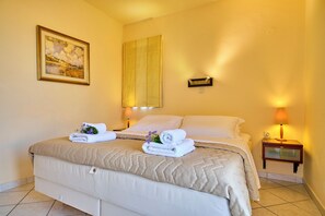 Una habitación de hotel con una cama, una mesita de noche con lámpara, un cuadro enmarcado en la pared y una ventana con cortina.