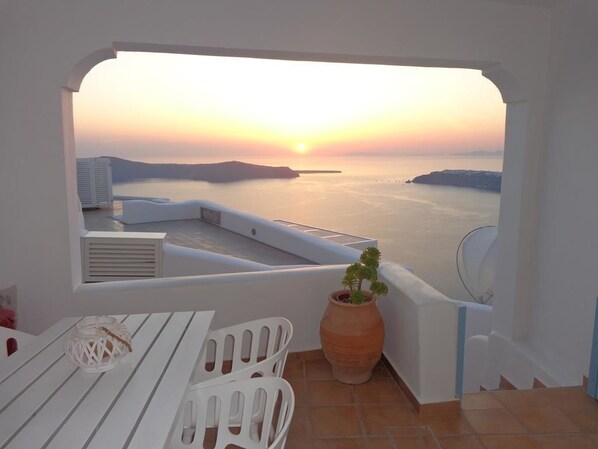 A terrace with a view of the sea at sunset, white furniture, and a potted plant. - Langas Villas (Santorini)