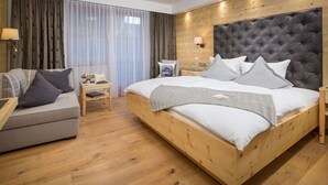 A modern hotel room with a wooden bed, a grey sofa, a wooden nightstand, and a large window with curtains. - Hotel Weinbauer (Schwangau)