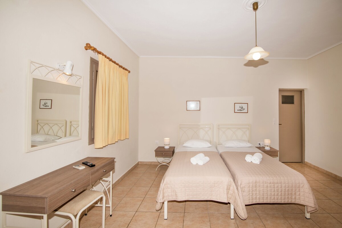 A bedroom with two beds, a desk, a chair, a bedside table with a lamp, and a mirror.