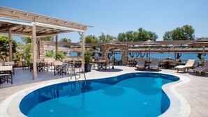 Une piscine entourée de chaises longues et de parasols, elle-même bordée d’une zone couverte avec des tables et des chaises.