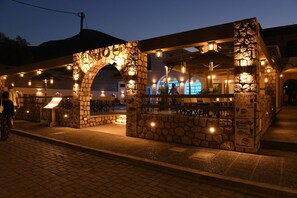 A stone-arched outdoor seating area with tables and chairs, illuminated by string lights. - Enjoy Villas (Santorini)