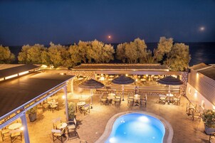An outdoor pool area with seating and umbrellas, surrounded by trees and overlooking the sea.
