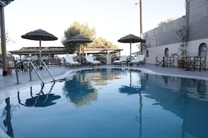 Piscina con sillas de descanso y sombrillas, rodeada de una zona de estar cubierta y un edificio con ventanales arqueados.