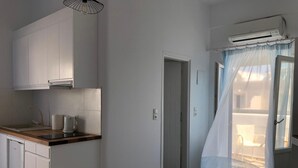 A compact hotel room with a kitchenette, a bed with a green bedspread, a bathroom with a shower curtain, and a view of the outside through a window with a sheer curtain. - Villa Gambas (Santorini)