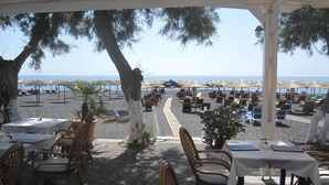 An outdoor dining area with tables and chairs, overlooking a beach with umbrellas and lounge chairs. - Villa Gambas (Santorini)