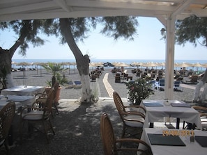 An outdoor dining area with tables and chairs, overlooking a beach with umbrellas and lounge chairs. - Villa Gambas (Santorini)
