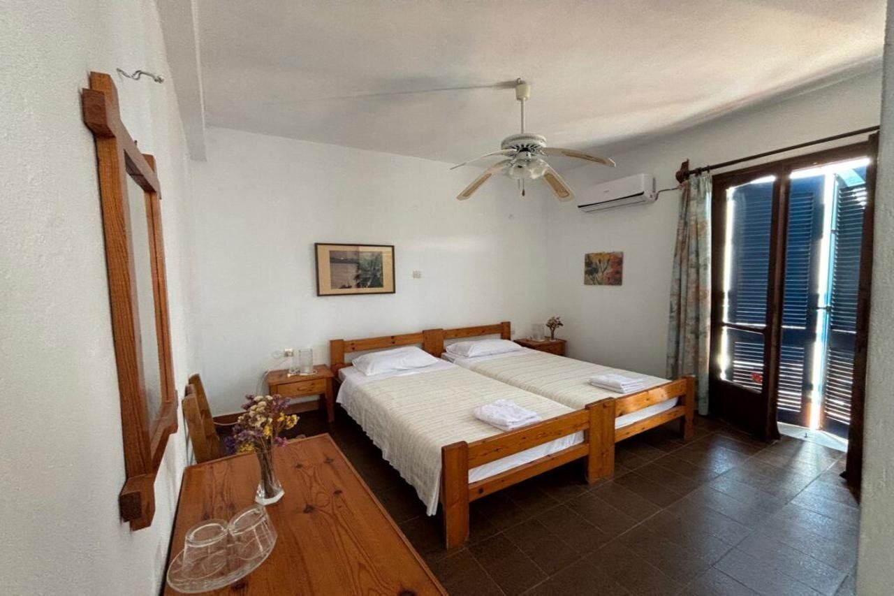 Un dormitorio con cama de madera, ventilador de techo, una mesa de madera con un jarrón y una ventana con contraventanas.