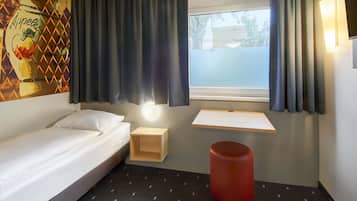 A hotel room with a bed, a small desk, a red stool, a wall-mounted shelf, a window with curtains, and a wall art piece.