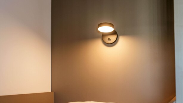 Un lit bien fait, avec une tête de lit adossée à un mur uni, un luminaire fixé au mur et un petit objet circulaire accroché au mur.