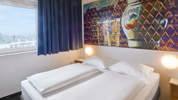 A hotel room with a bed, two bedside tables, a window with curtains, and a wall mural.